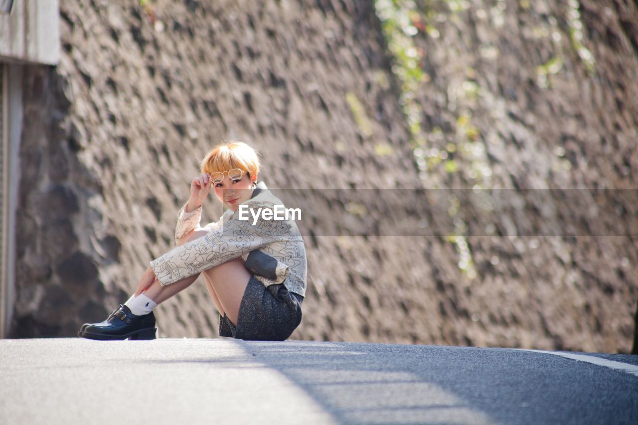 Full length portrait of young woman sitting on road during sunny day