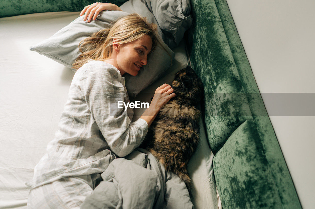 A woman in the morning after waking up lies in bed and pets a domestic cat.
