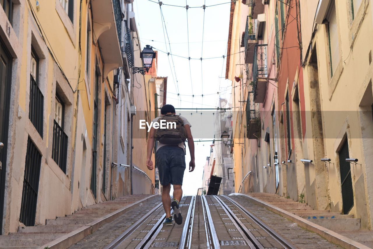 REAR VIEW OF MAN WALKING ON RAILROAD TRACK AMIDST BUILDINGS