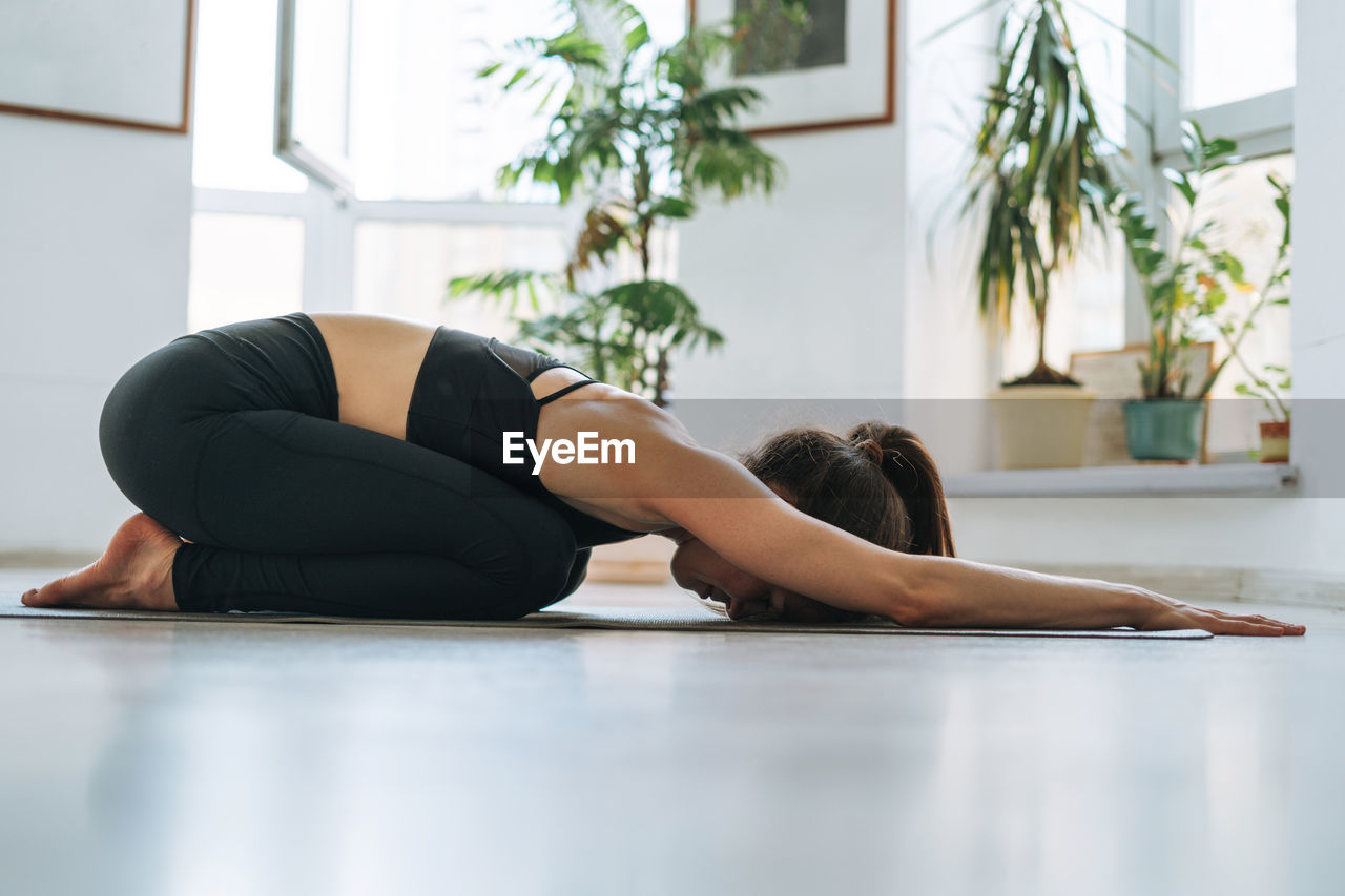 Young fit woman practice yoga doing asana in light yoga studio with green house plant