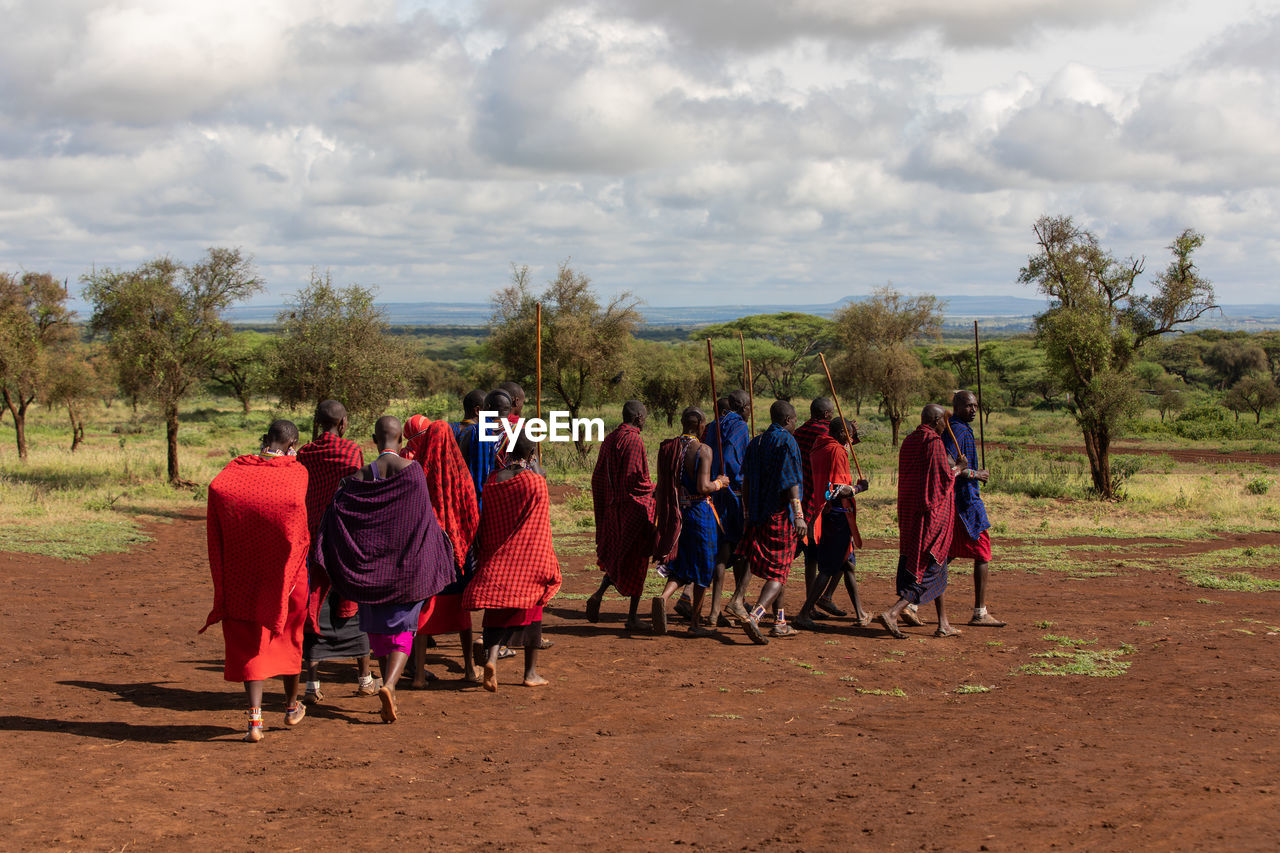 REAR VIEW OF PEOPLE WALKING ON FIELD BY TREES AGAINST SKY