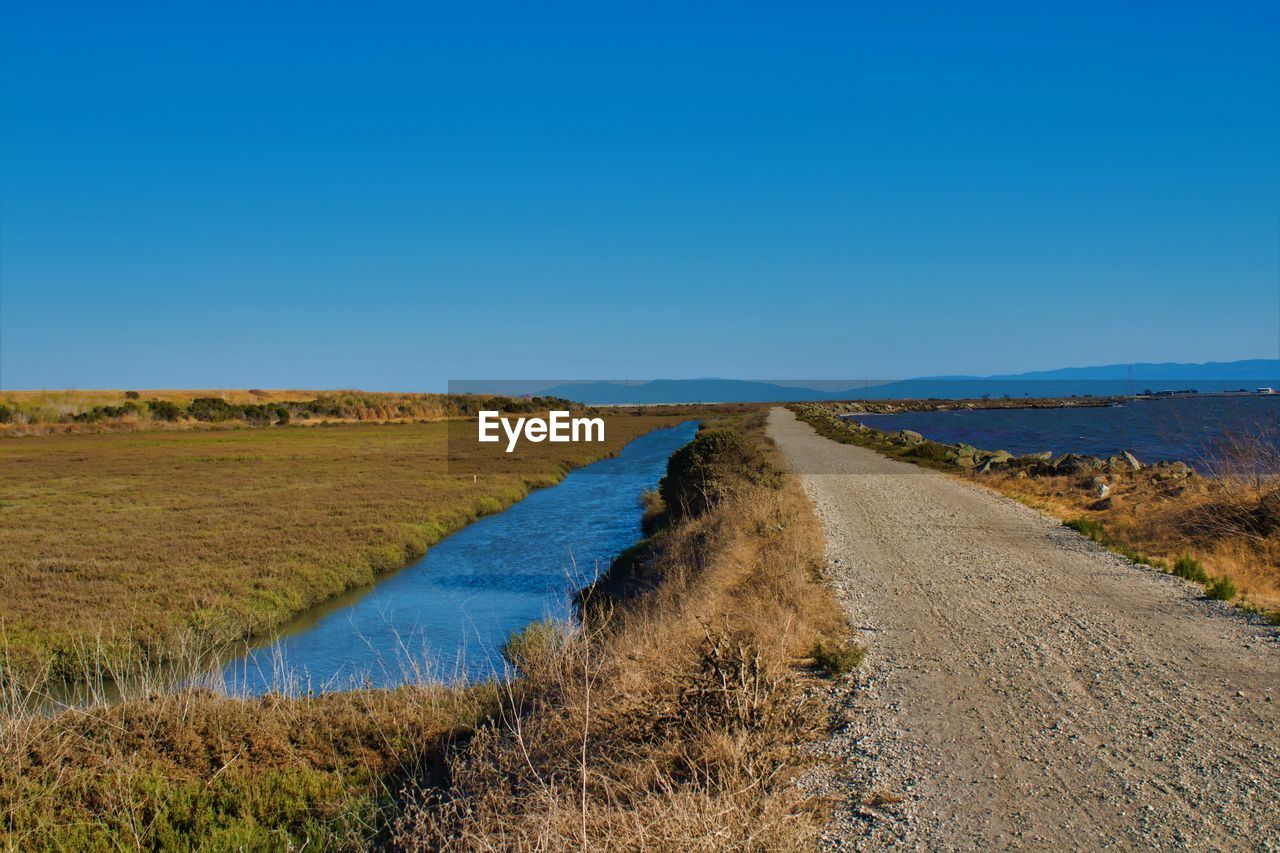 Road amidst field against clear blue sky