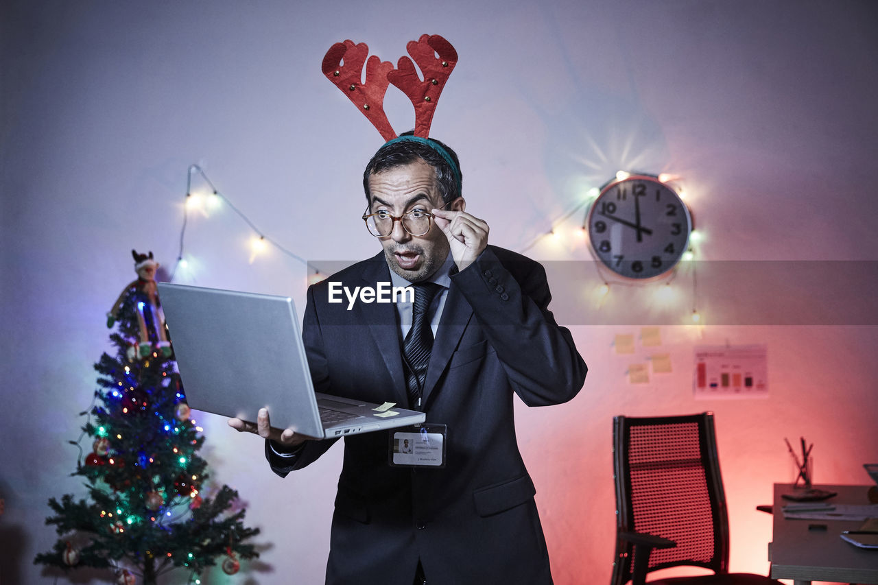 Shocked businessman wearing headband holding laptop against illuminated christmas tree and lights at home