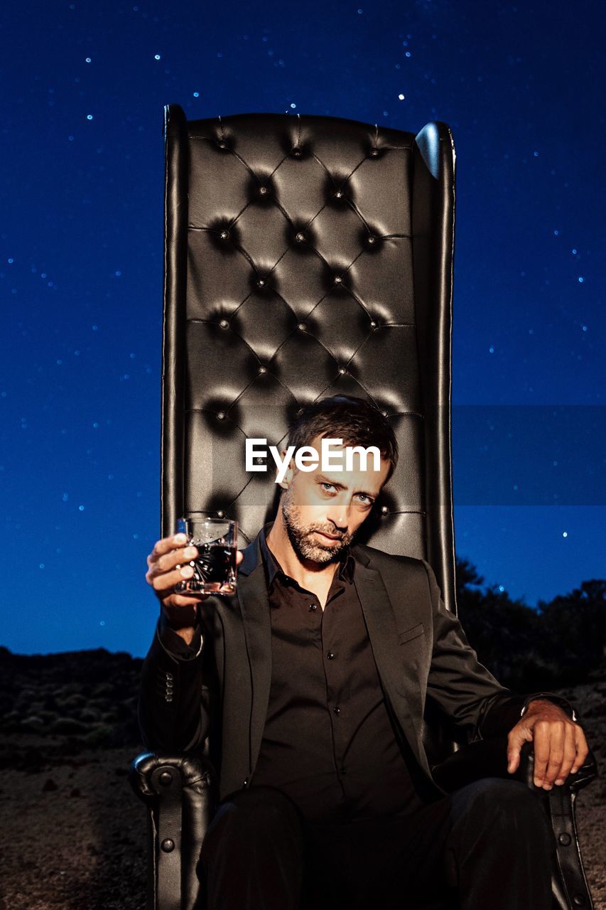 Portrait of man holding whiskey glass while sitting on sofa at night