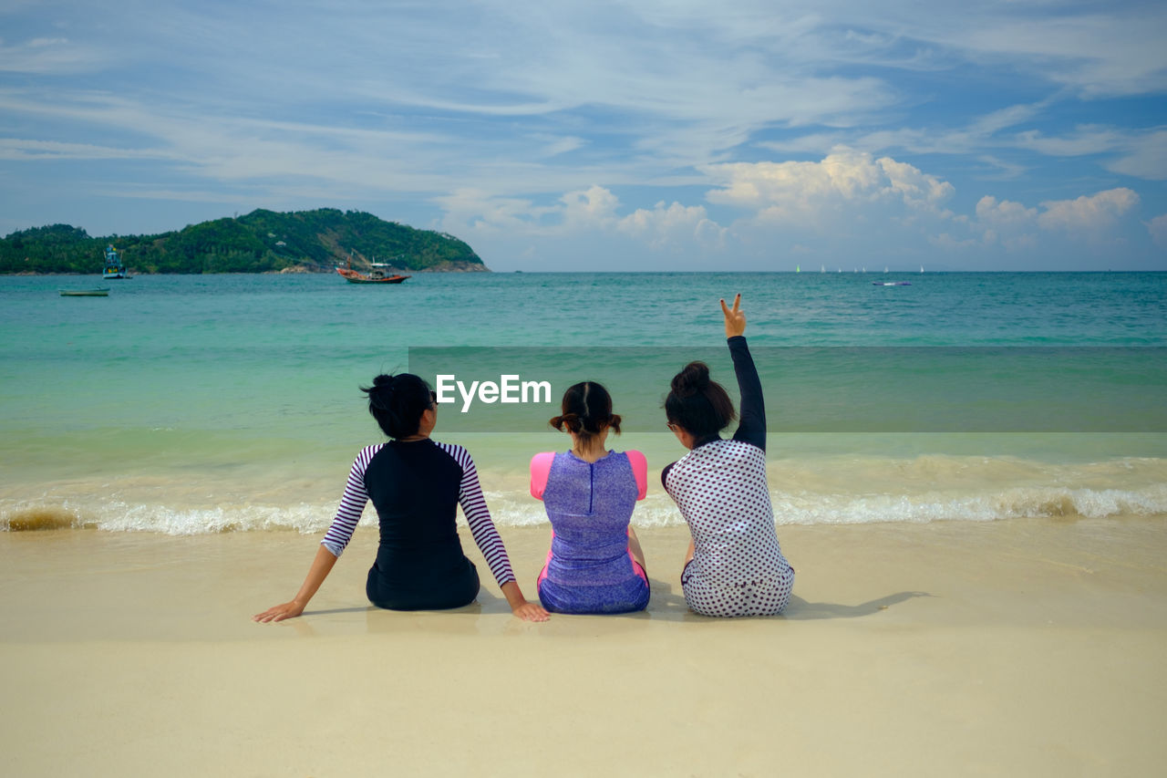 Rear view of female friends sitting on shore at beach against sky