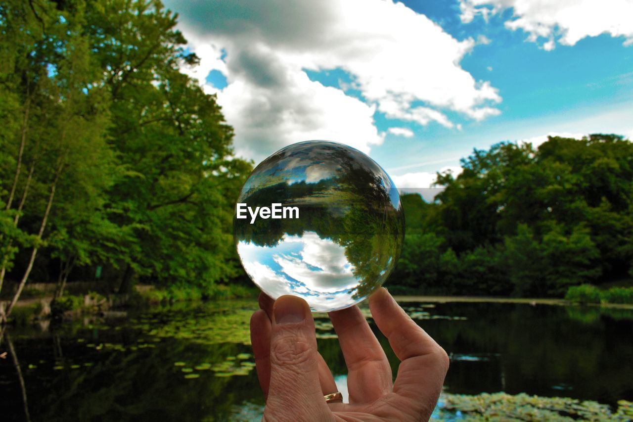 Close-up of hand holding crystal ball with reflection of trees, lake and sky in dulwich park, london