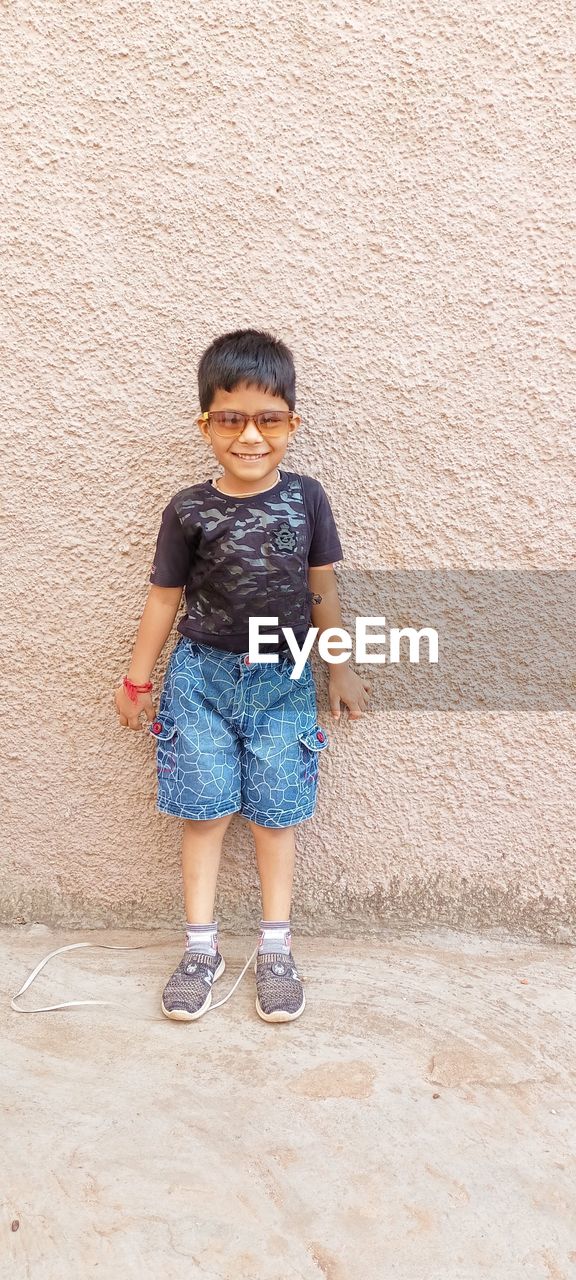 full length, childhood, child, one person, casual clothing, blue, men, front view, standing, portrait, wall - building feature, toddler, footwear, spring, architecture, clothing, day, looking at camera, person, innocence, smiling, shoe, leisure activity, pattern, lifestyles, looking, outdoors, cute, shorts, happiness, copy space, emotion, photo shoot