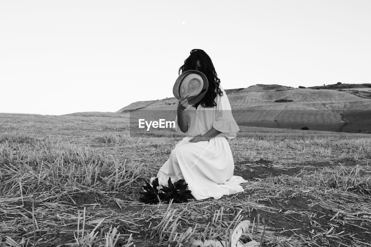 Woman covering face with hat on land against clear sky