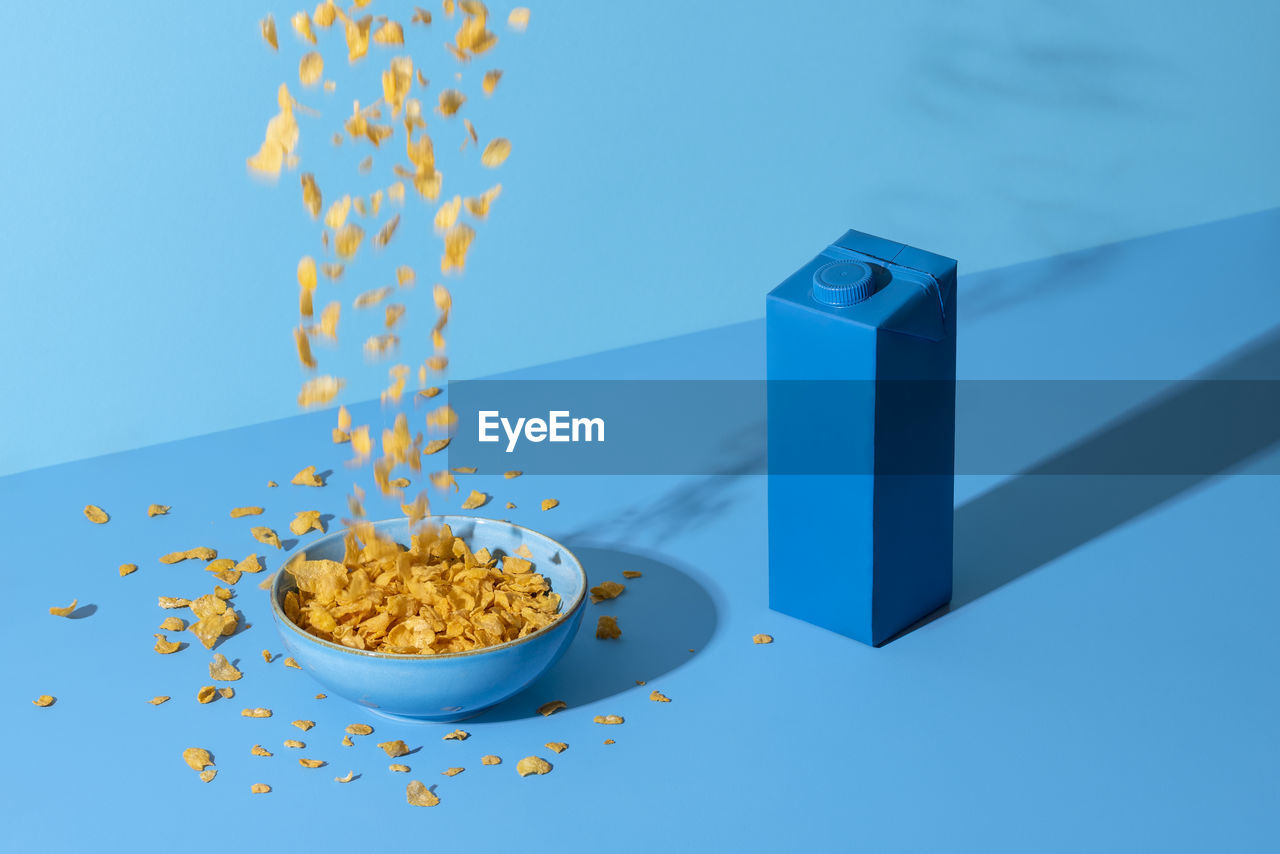 Pouring cereals in a bowl on blue table. breakfast table, cornflakes cereals in bowl and a milk box