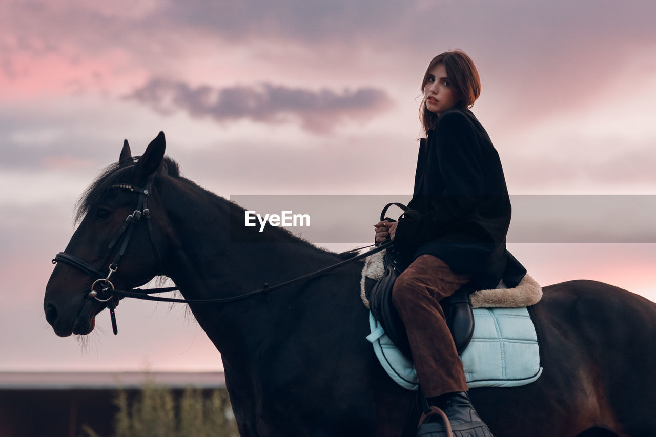 side view of young woman riding horse against sky