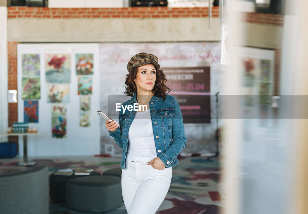 Charming young woman in jeans jacket using mobile phone in modern art gallery exhibition