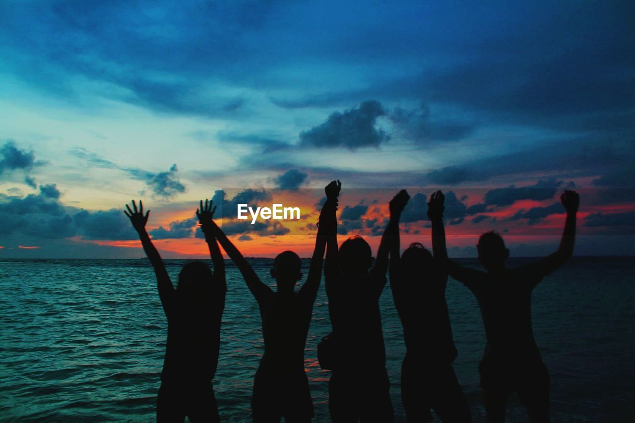 Silhouette people with arms raised standing in sea during sunset