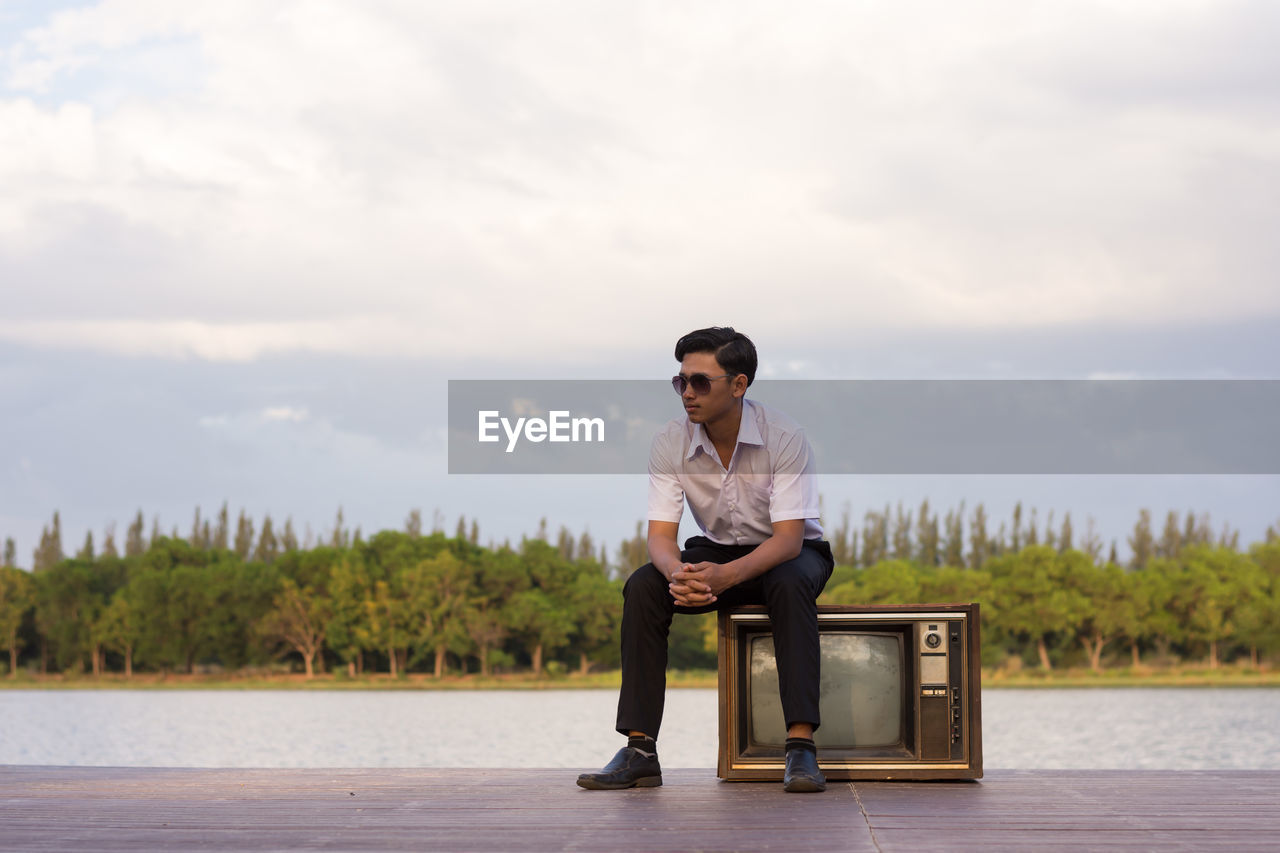 Teenage boy sitting on television set at lakeshore against sky