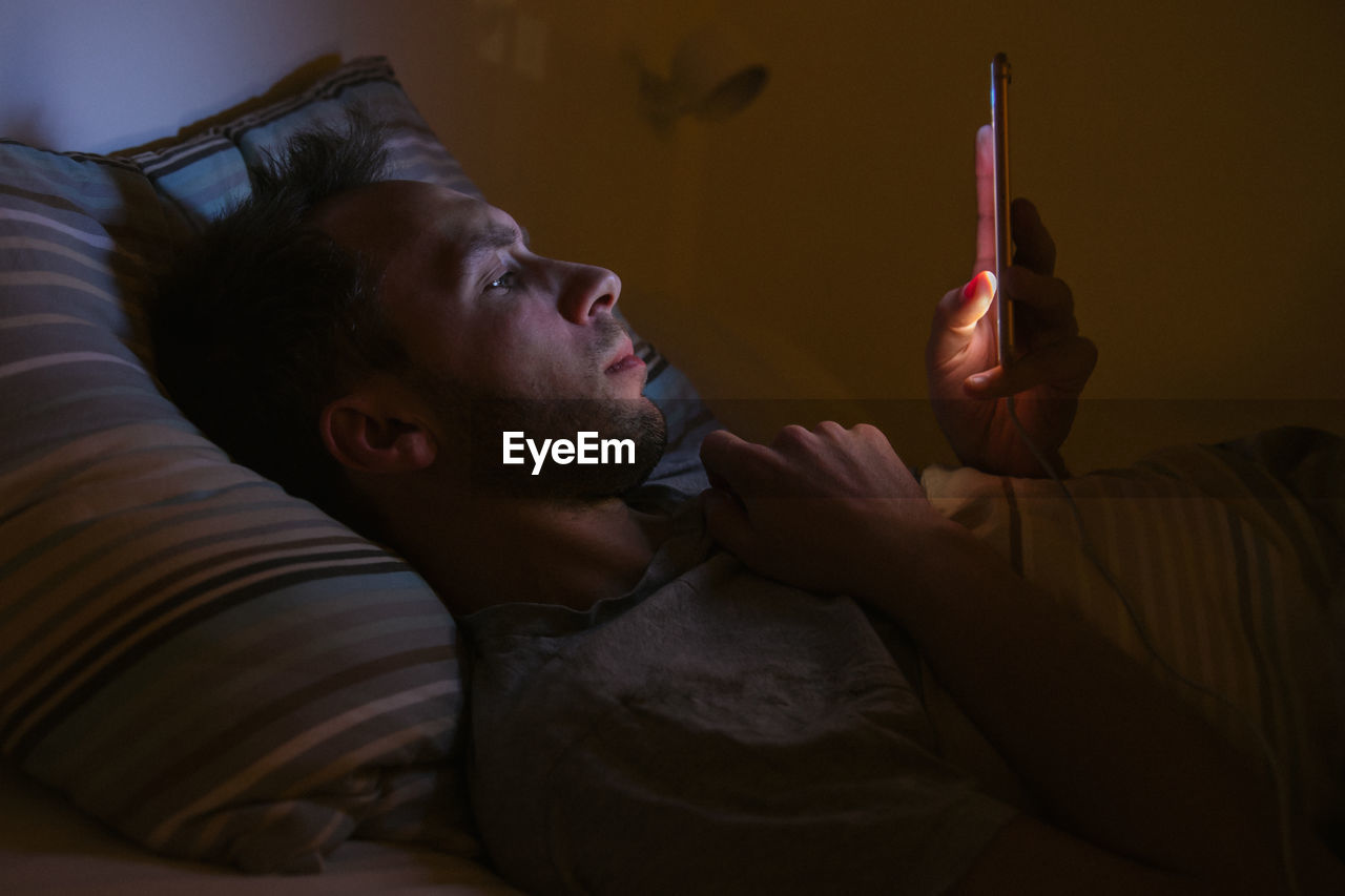 Side view of man using smart phone while lying on bed at home