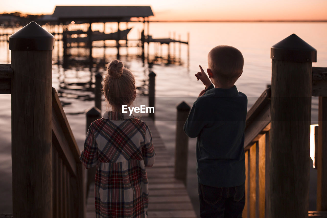 Rear view of brother pointing towards lake while standing by sister during sunset