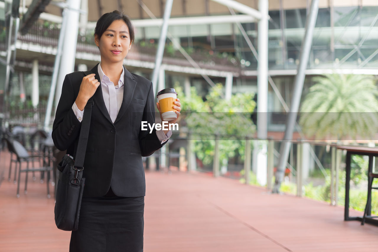 Businesswoman holding disposable cup while standing against building in city