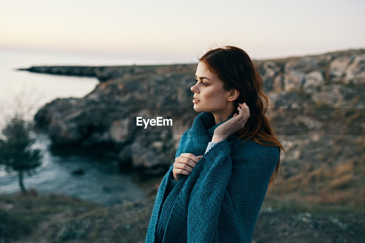 YOUNG WOMAN LOOKING AWAY WHILE STANDING ON ROCK AT BEACH