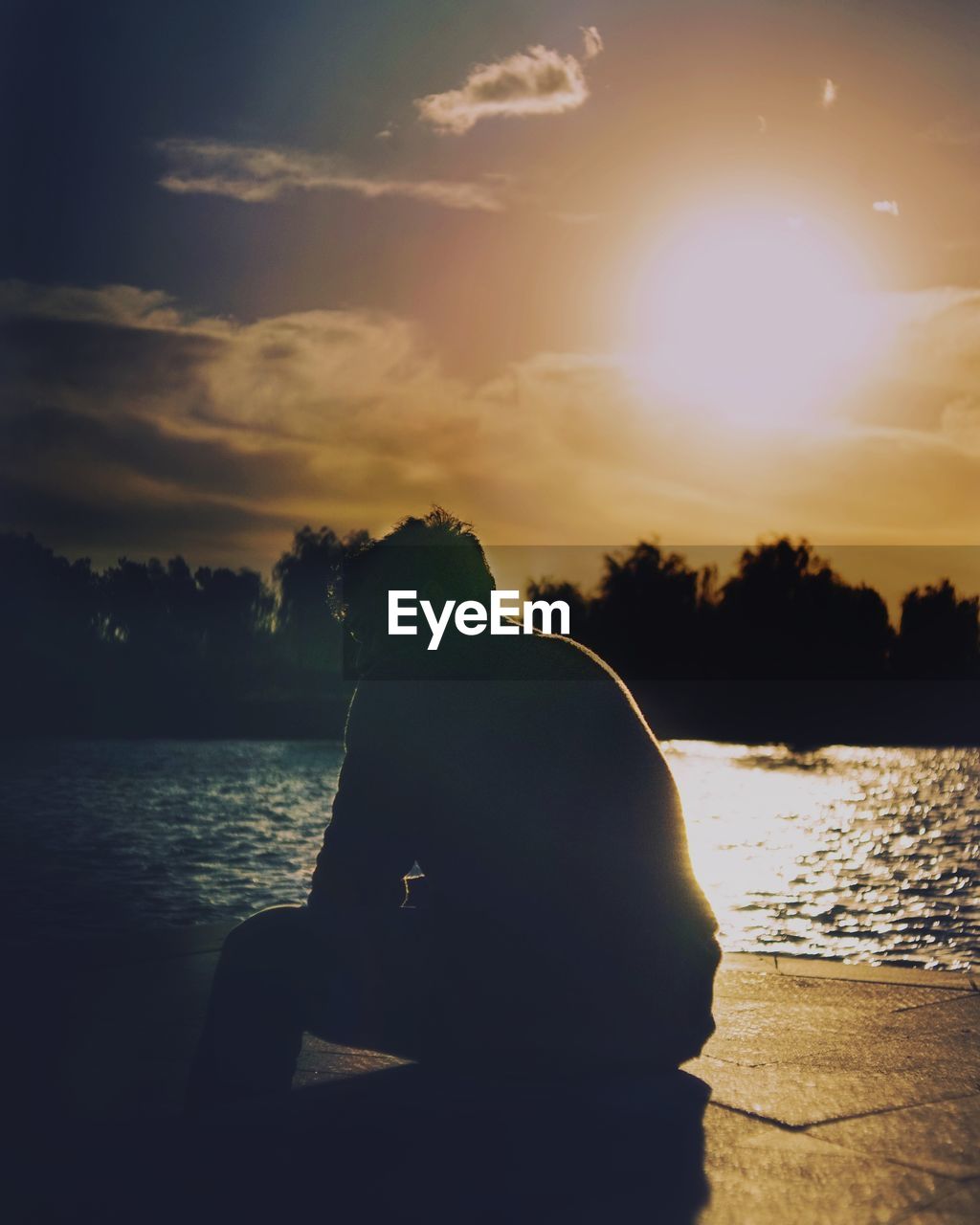 REAR VIEW OF WOMAN SITTING BY LAKE AGAINST SUNSET SKY