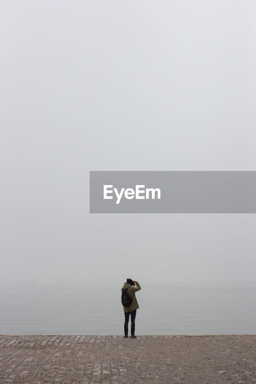 REAR VIEW OF MAN STANDING ON SEA SHORE AGAINST SKY