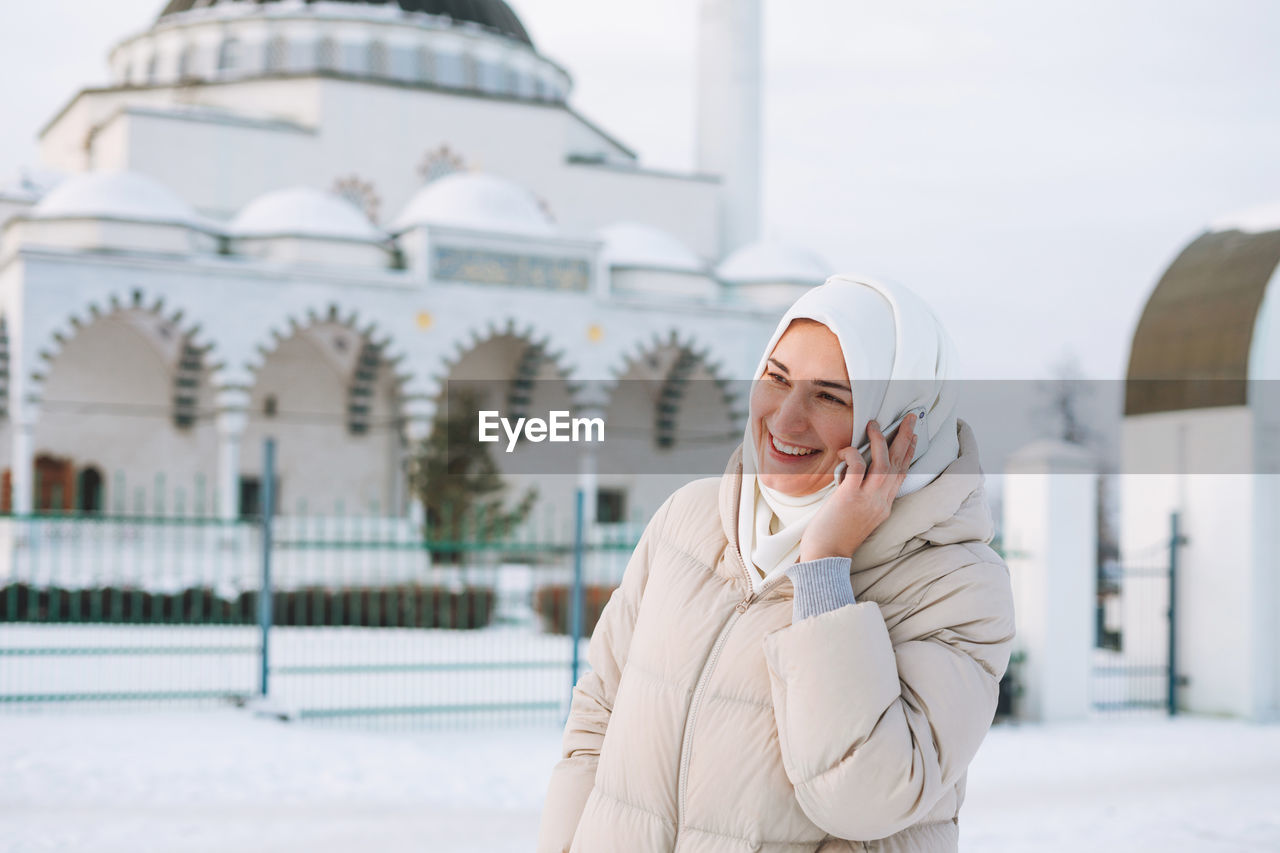 Beautiful smiling young muslim woman in headscarf using mobile against mosque in winter season