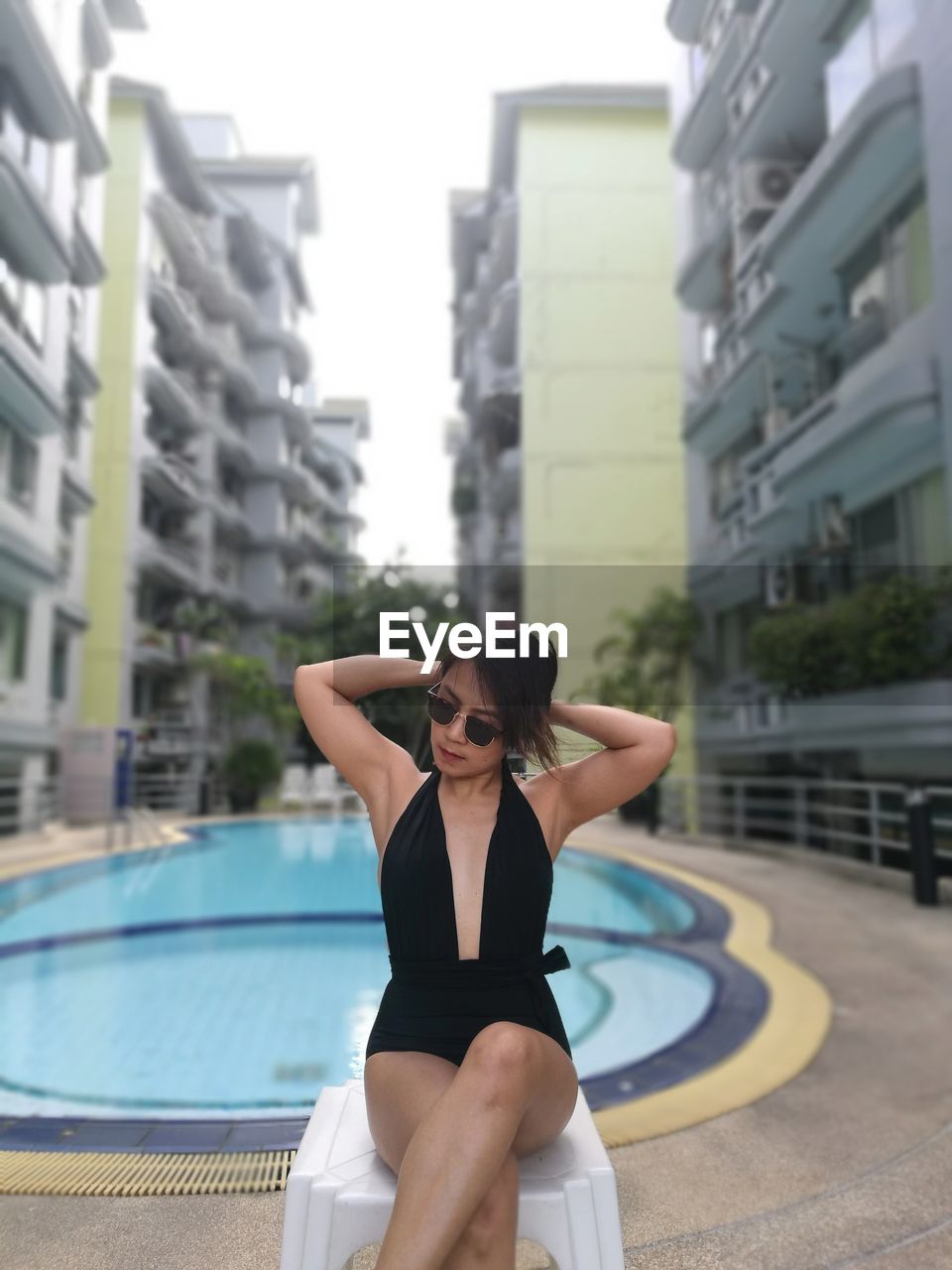 Woman in swimwear sitting on stool by swimming pool amidst buildings