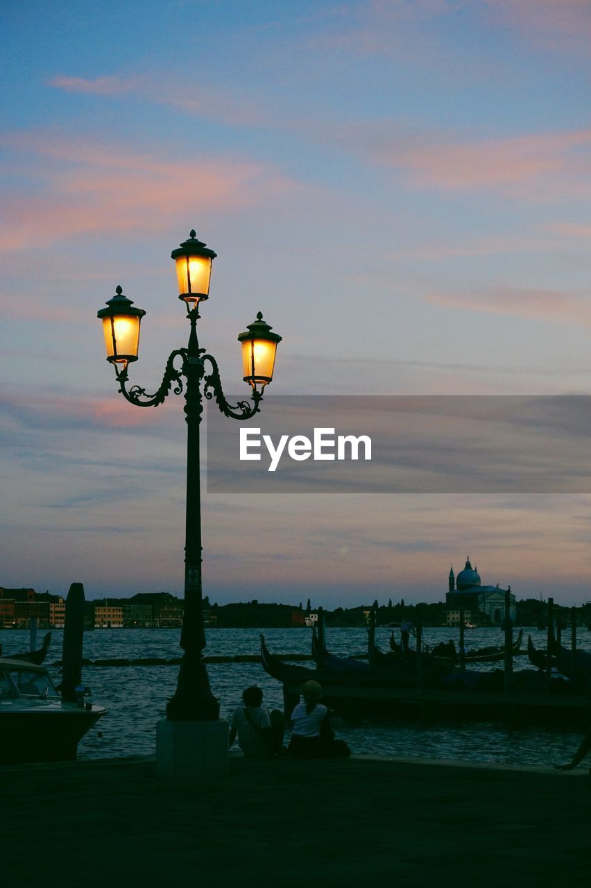 Illuminated street light by river against sky during sunset