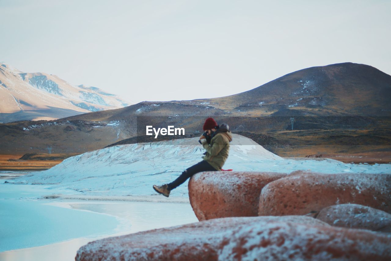 Side view of man photographing while sitting on rock against sky