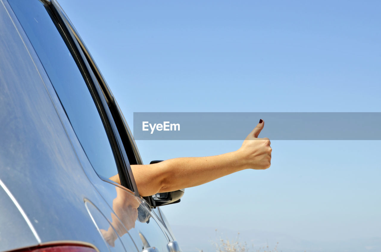 Cropped hand of woman gesturing in car window against blue sky