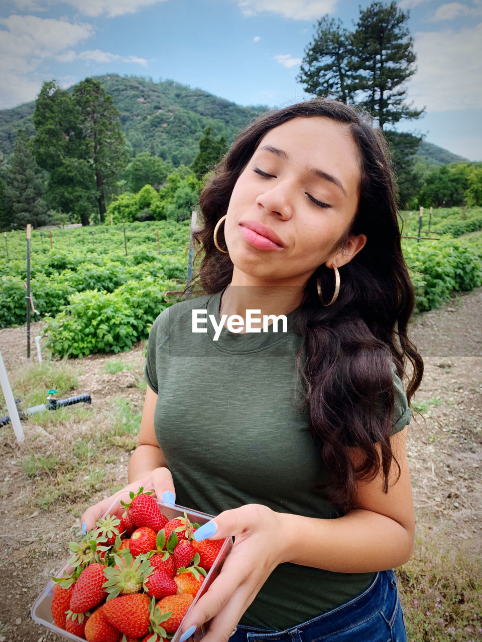 Girl with eyes closed holding strawberries in container while standing against sky