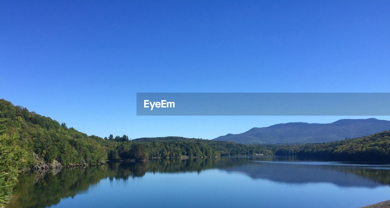 Scenic view of calm lake by mountain against clear blue sky