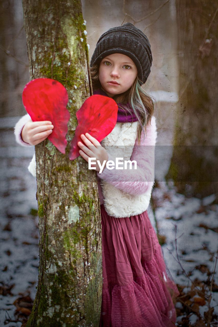 Portrait of cute girl holding heart shape standing by tree trunk