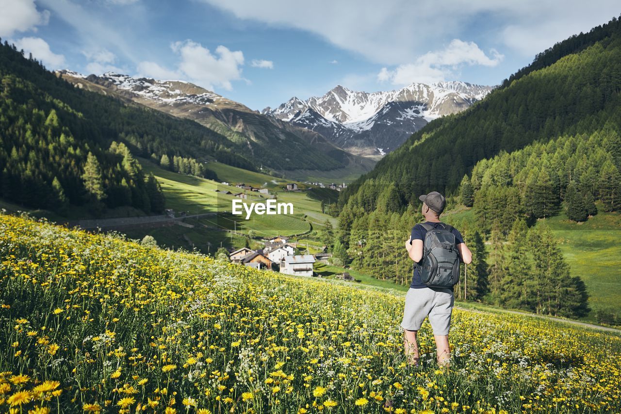 Rear view of man with backpack standing amidst flowering plants on field against mountains