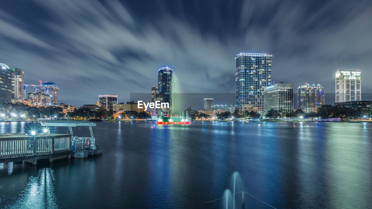 Illuminated city at waterfront against cloudy sky