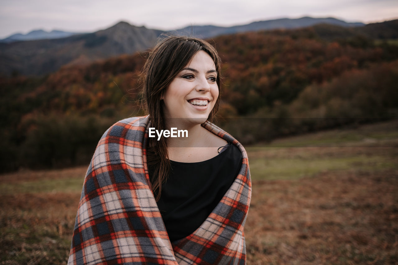 Delighted female wrapped in cozy checkered blanket standing on hill in highlands and looking away