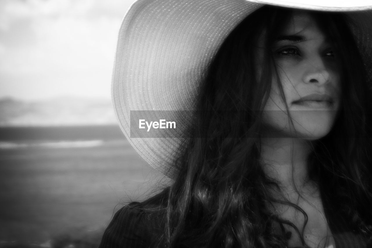 Close-up of young woman wearing hat while looking away at beach against sky