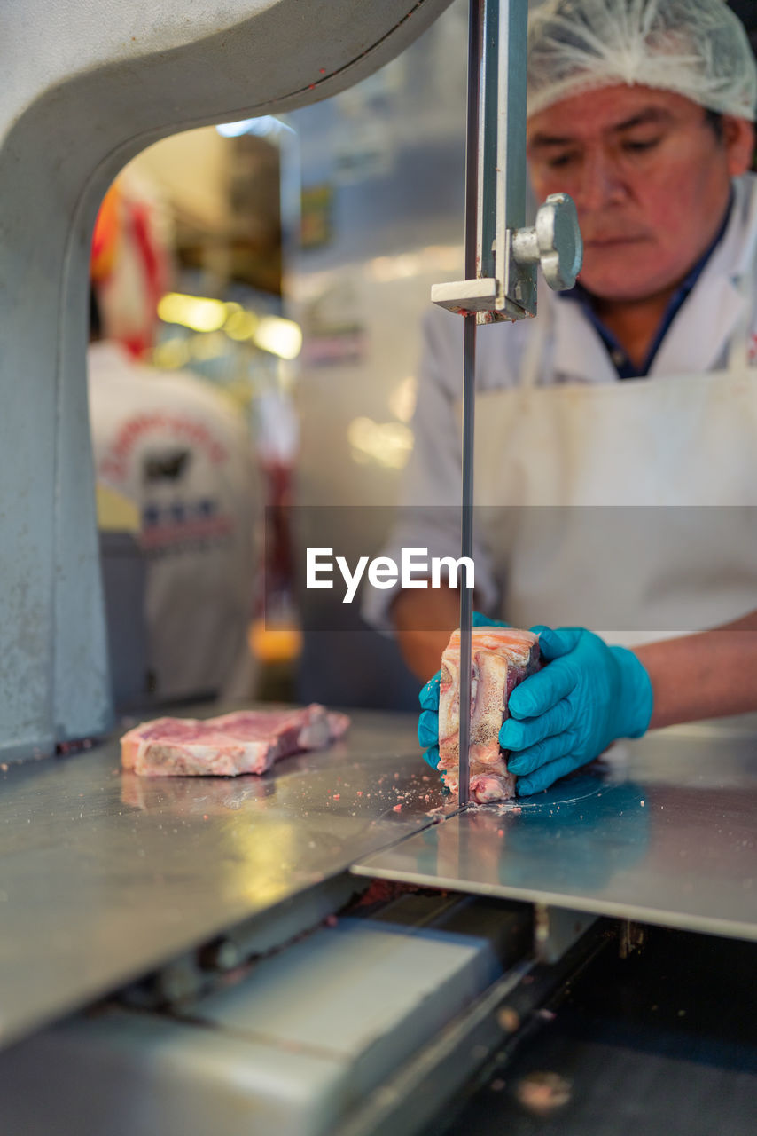 Mature man in apron using electric tabletop saw to slice piece of frozen meat during work in butchery