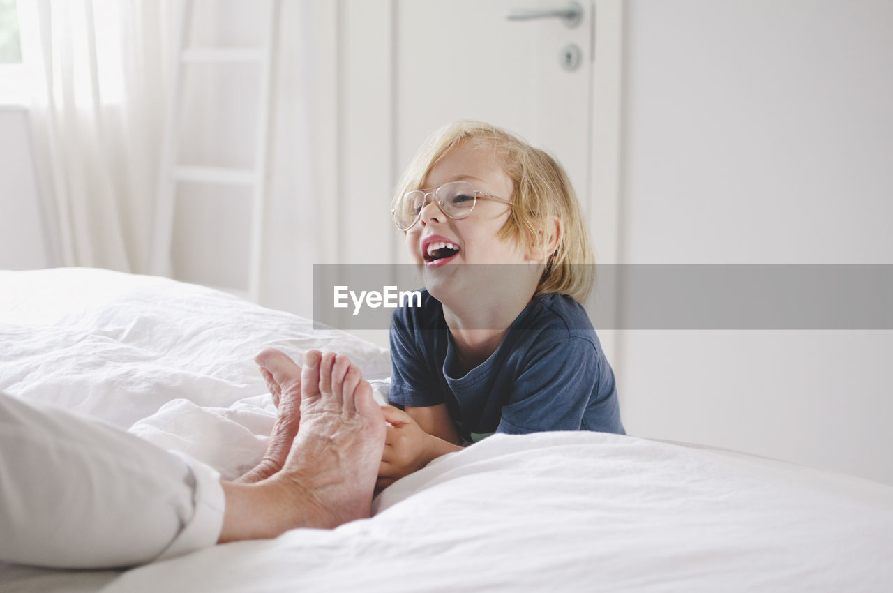 Happy grandson tickling grandmother's bare feet on bed at home