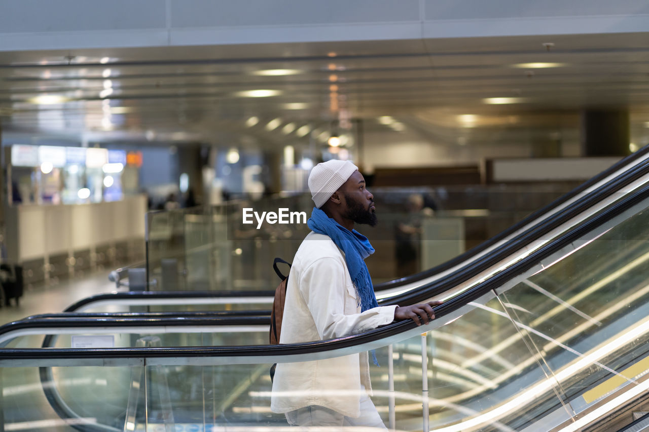 African american passenger man with suitcase stands on escalator, holds handrail in airport terminal