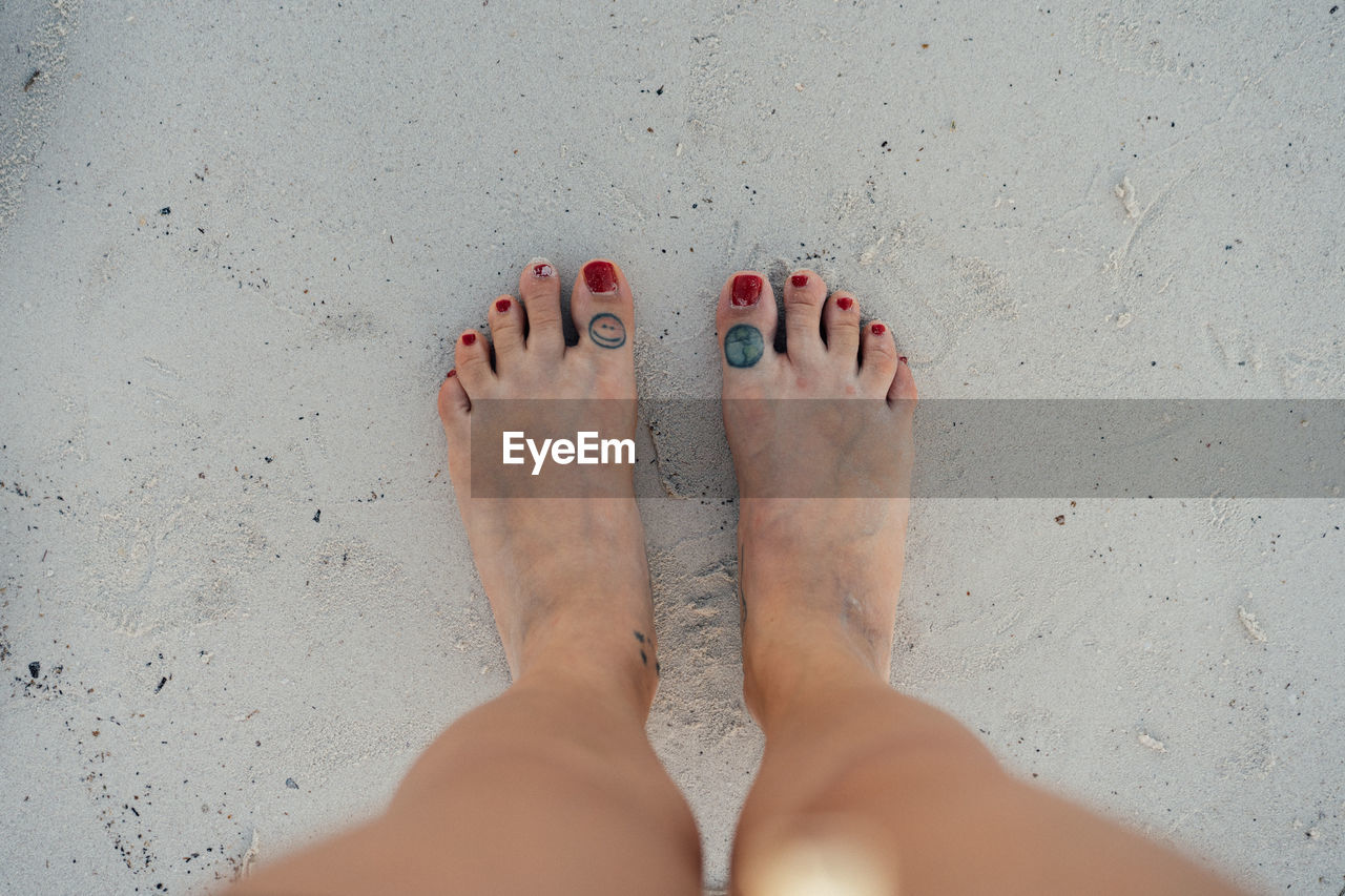 Womans feet with red nails and tattoos on white sand.