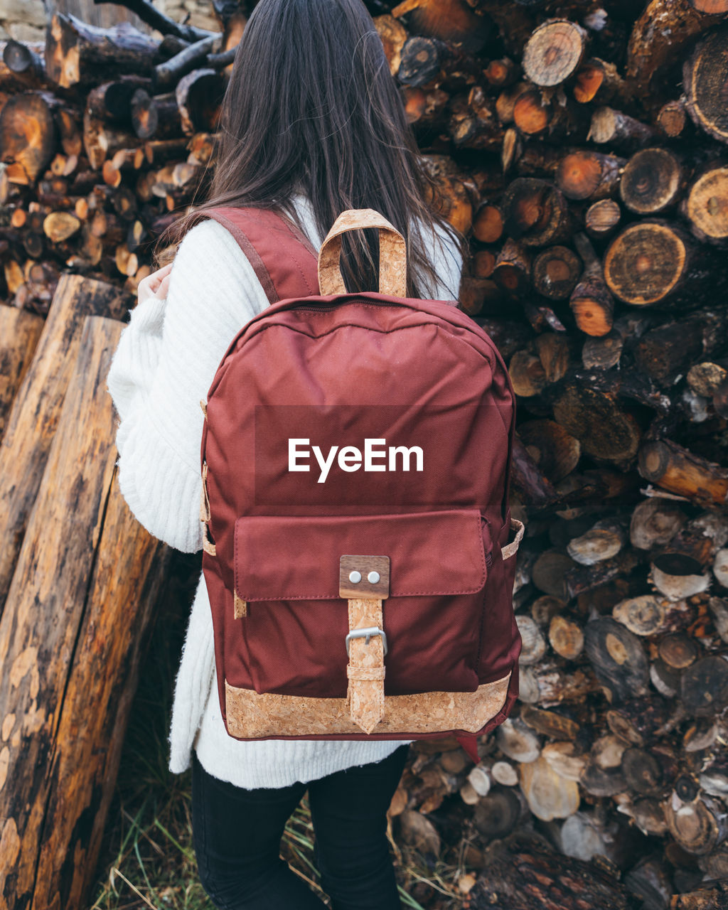 Back view of unrecognizable active young female in white sweater with stylish red backpack standing near stacked wooden logs in autumn day in countryside