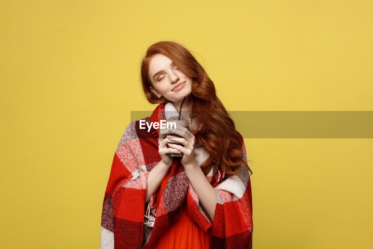 Young woman wrapped in shawl having drink while standing against yellow background