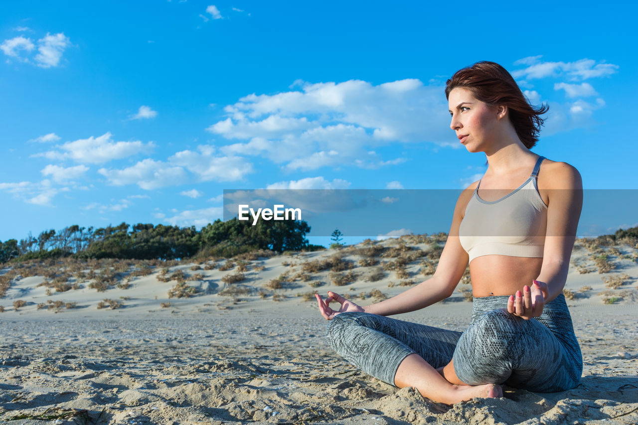 Young woman doing yoga at beach against cloudy sky