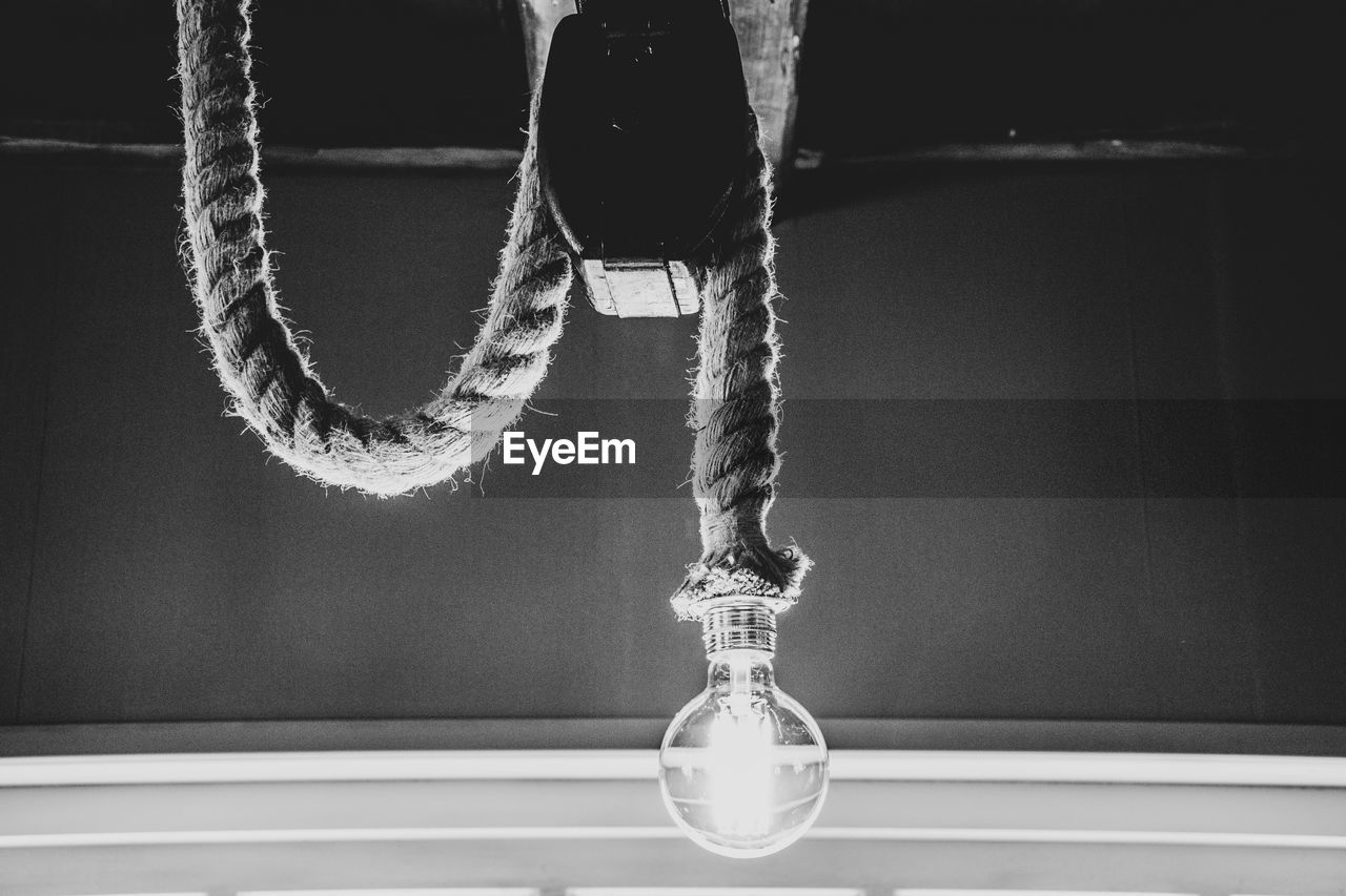 Close-up of illuminated bulb hanging from rope against wall