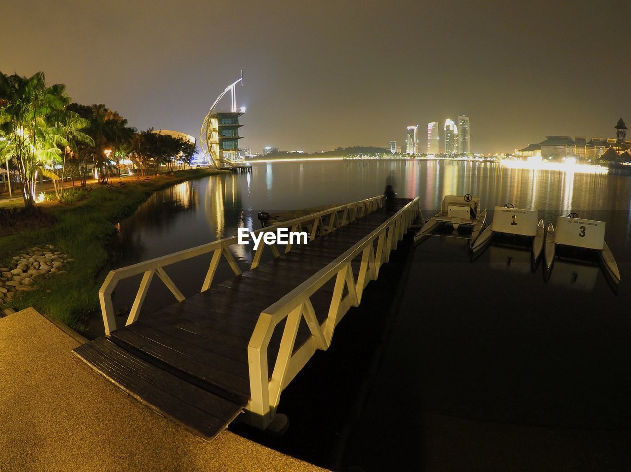 Pier by calm lake in illuminated city against clear sky at night