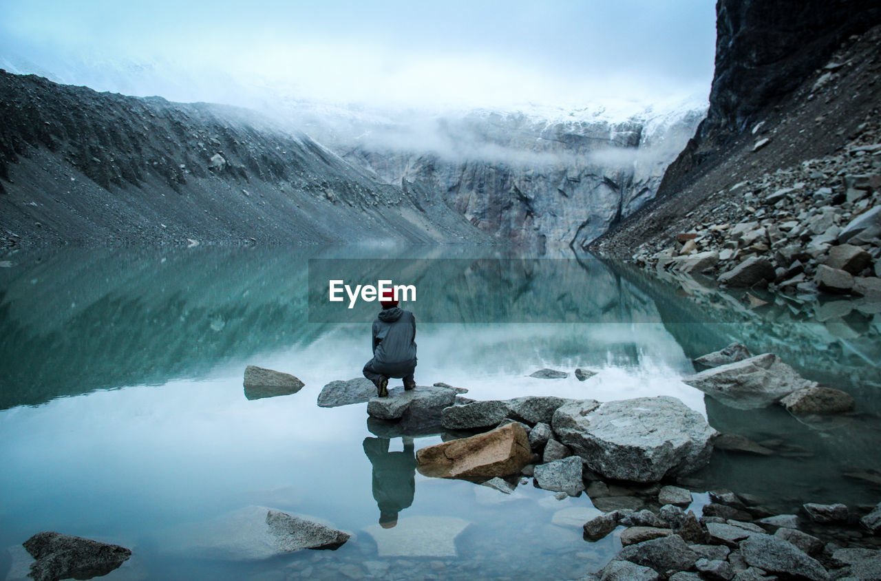 Woman sitting on rock against mountains in lake during winter