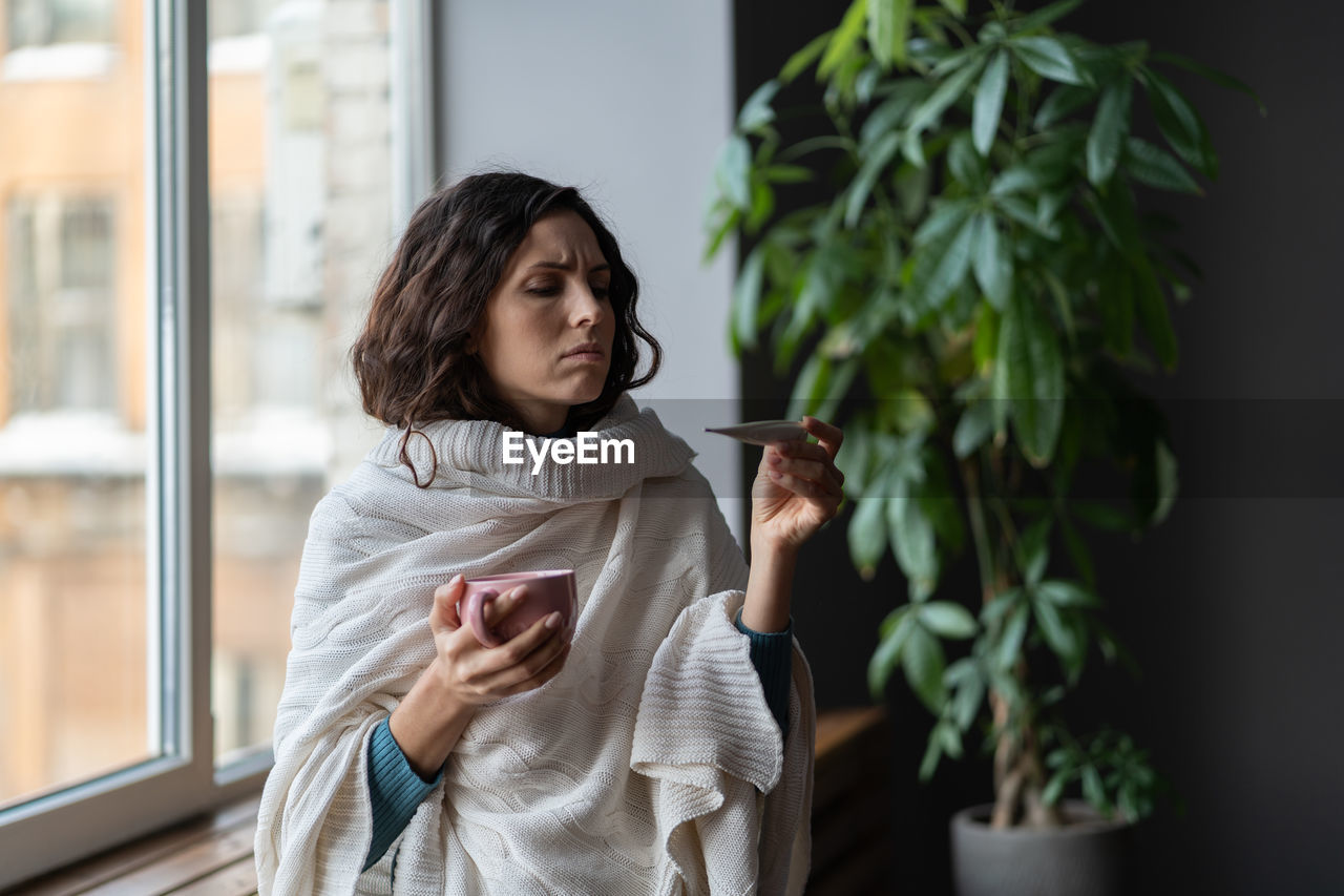 Sick unhealthy young woman with high fever looking at thermometer, checking body temperature