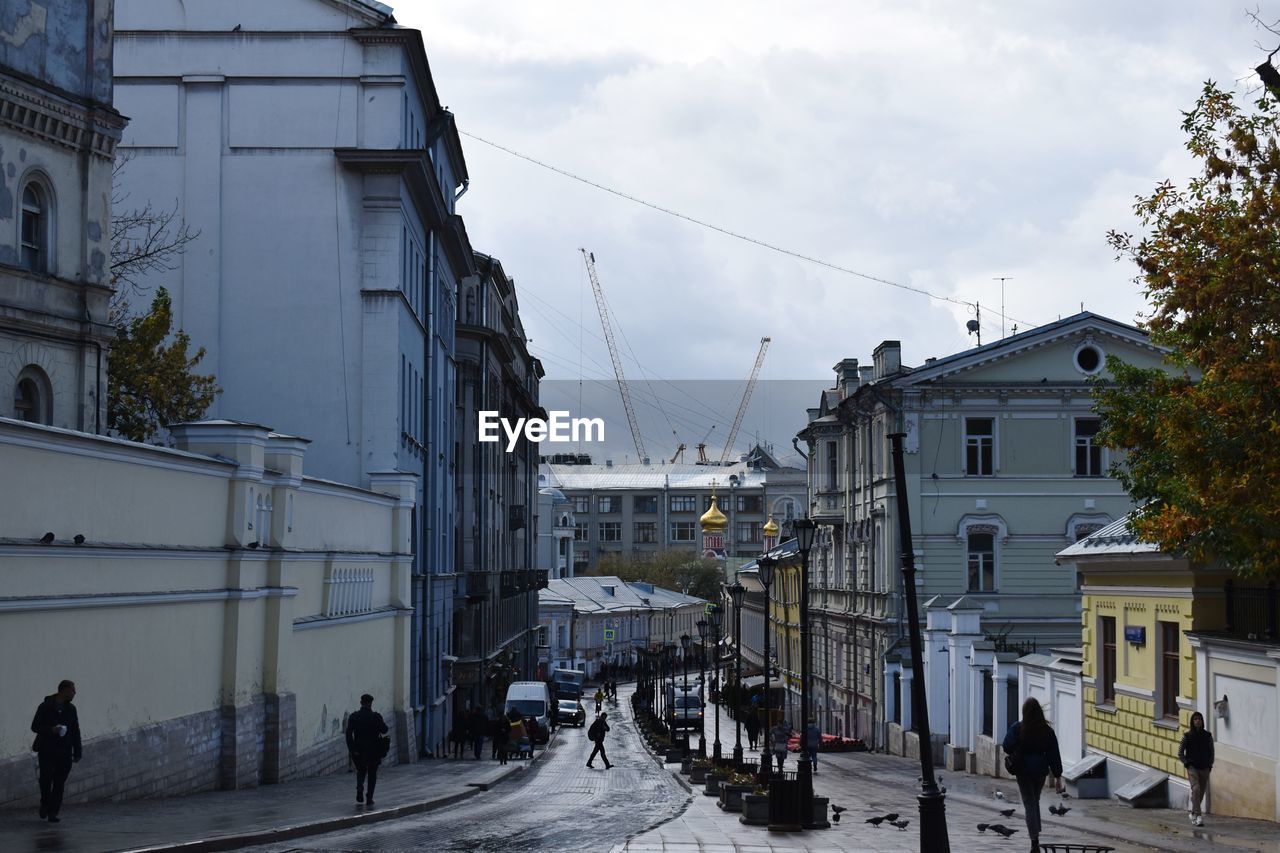 PEOPLE WALKING ON ROAD AMIDST BUILDINGS AGAINST SKY