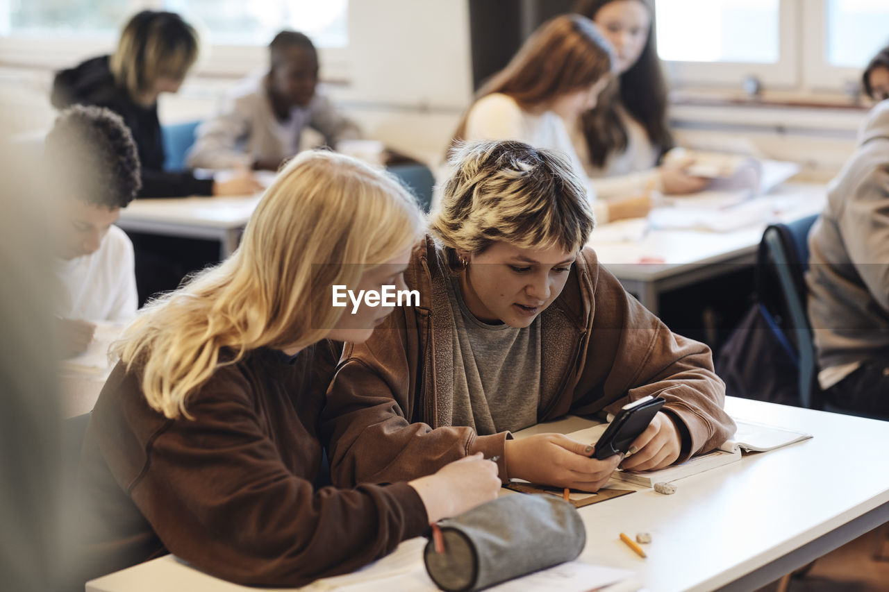 Teenage girl sharing smart phone with blond female friend sitting at desk in classroom