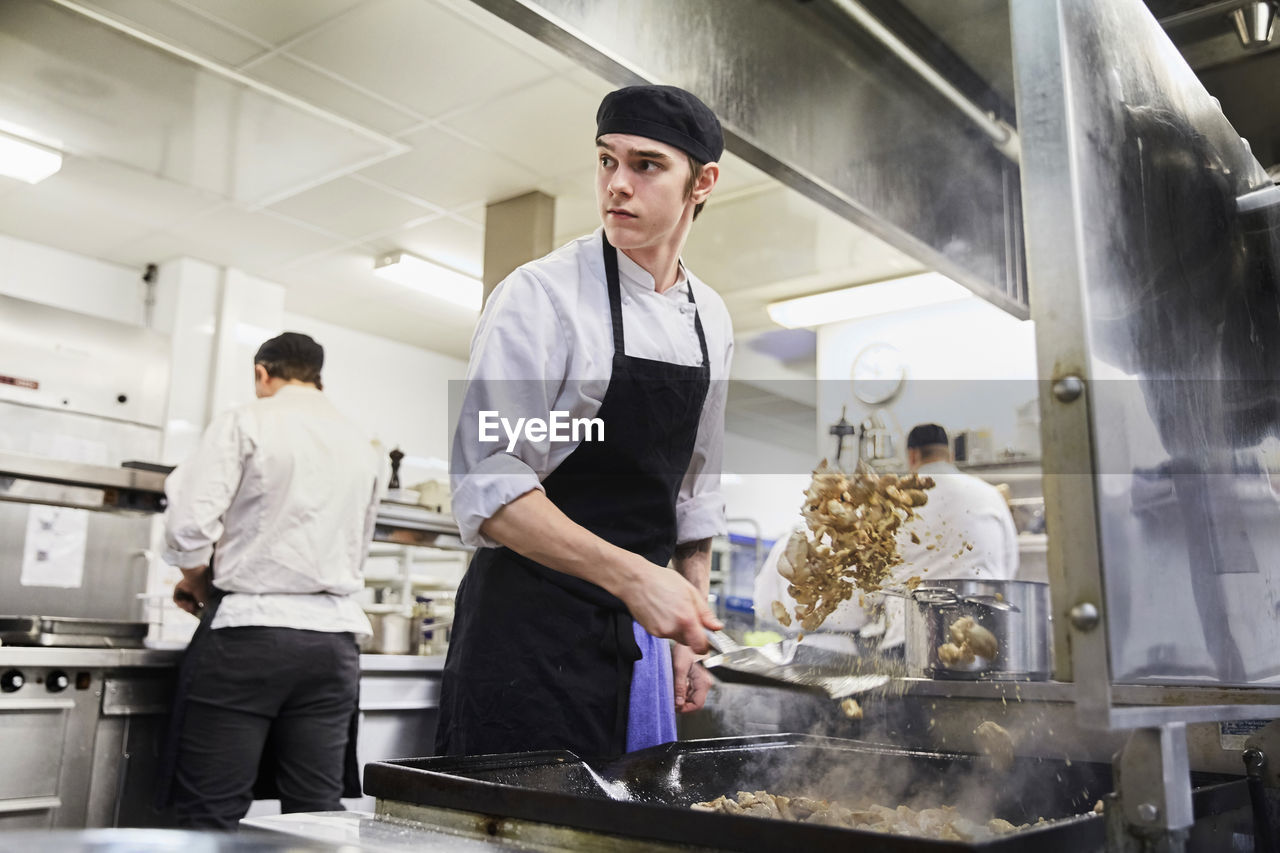 Male chef student tossing meat with colleagues in background at cooking school