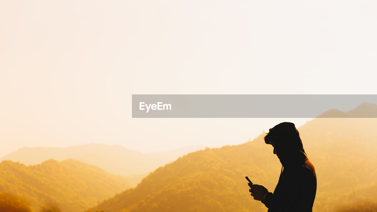 Side view of man using phone by mountain against clear sky